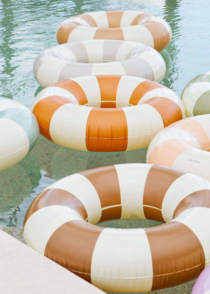 Petites Pommes Charleston Pool Float floating in pool with other floats. It is a striped pool ring in a rich brown and cream for adults.