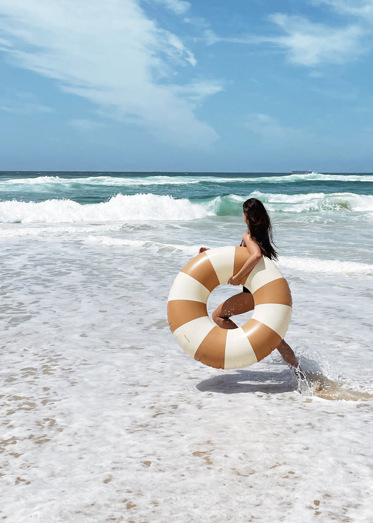 Petites Pommes Dolce Pool Float being held by an adult woman to show size. It is a striped pool ring in caramel brown and cream for adults.
