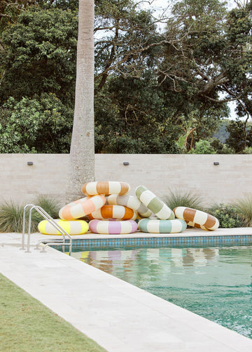 Petites Pommes Dolce Pool Float stacked on side of pool with other floats. It is a striped pool ring in caramel brown and cream for adults.