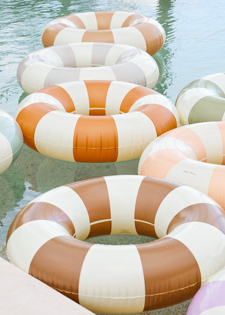 Petites Pommes Tangerine Pool Float floating in pool with other floats. It is a striped pool ring in burnt orange and cream for adults.